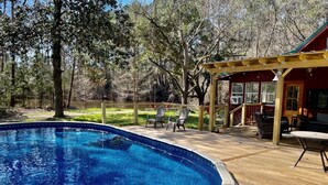 Una piscina con cubierta de madera, rodeada de árboles y un patio techado con mobiliario exterior.