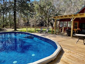 Una piscina con cubierta de madera, rodeada de árboles y un patio techado con mobiliario exterior.