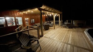 Una terraza de madera con asientos exteriores, un espacio cubierto con luces colgantes y una piscina.
