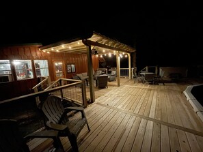 Una terraza de madera con asientos exteriores, un espacio cubierto con luces colgantes y una piscina.