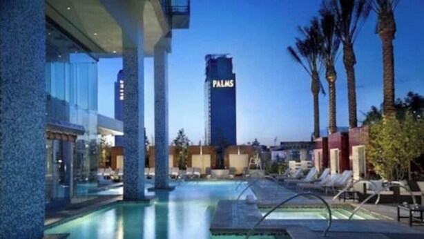 Un área de piscina con sillones de descanso, un edificio alto con la leyenda "PALMS" al fondo y palmeras.