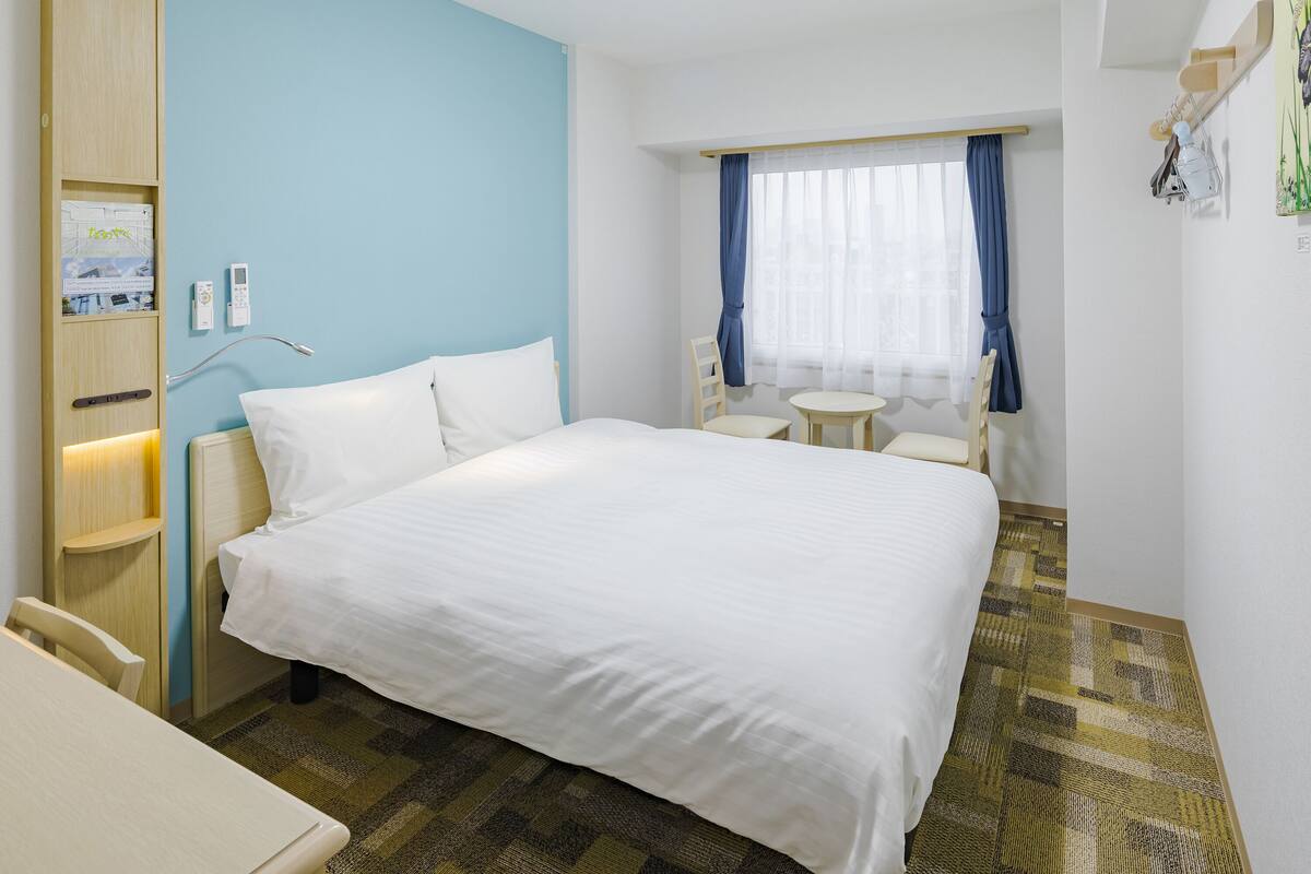 Ein Hotelzimmer mit einem Bett, Nachttischen, einem kleinen runden Tisch, zwei Stühlen, einem Fenster mit Vorhängen und einem Wandregal mit Lampe und kleiner Pflanze.