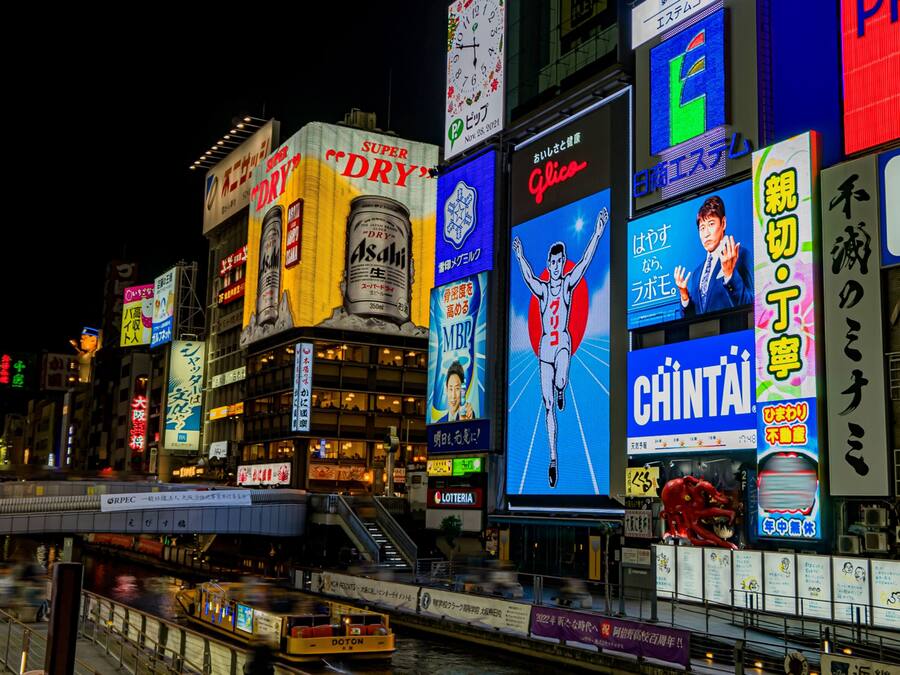 Una calle urbana concurrida de noche, con vallas publicitarias iluminadas y señalizaciones en un idioma extranjero.
