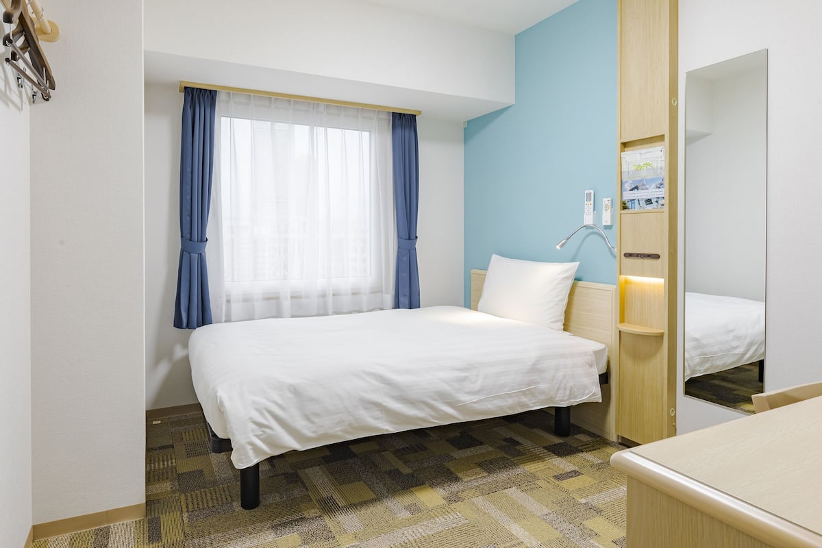 Ein Hotelzimmer mit einem Bett, einem Nachttisch, einem Spiegel, einem Fenster mit Vorhängen und einer wandmontierten Bedienkonsole.