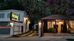 Entrada de un hotel con letrero, acceso mediante portón y una pequeña zona cubierta con asientos.