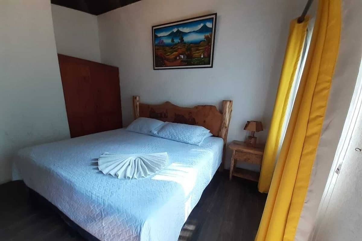 A bedroom with a bed, a wooden headboard, a nightstand, a lamp, a painting on the wall, and a yellow curtain.