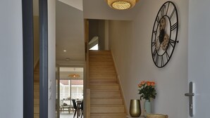 Un couloir avec un escalier en bois, un luminaire en fibres tressées, une horloge murale et une petite console ornée d’objets décoratifs.
