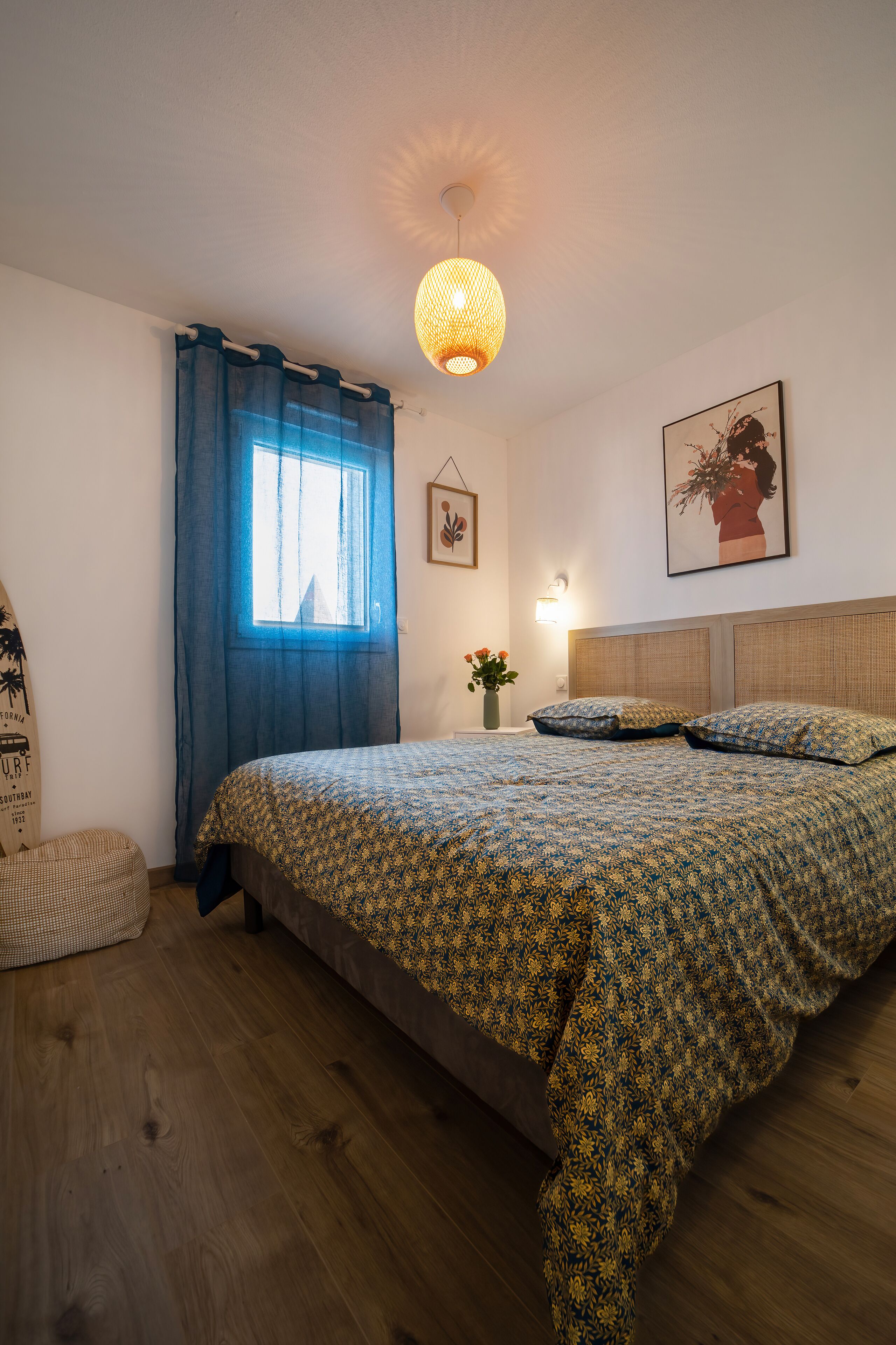 A bedroom with a bed, a window with curtains, a wall-mounted light, a framed picture, and a small table with a vase.