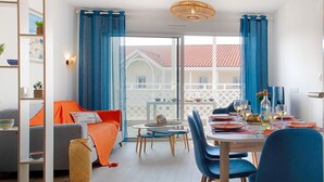 Un salon moderne avec un coin repas, comprenant un canapé, une table à manger, des chaises et une vue sur un immeuble à travers de grandes fenêtres.