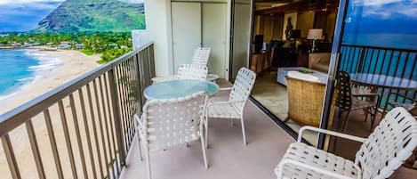 Un balcón con puerta de cristal que da a una habitación, mobiliario de mimbre blanco y vista a la playa y la montaña.