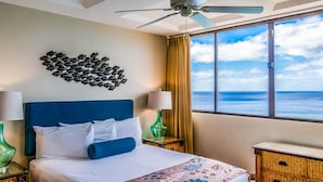 A hotel room with a bed, a ceiling fan, a view of the ocean, and a decorative wall piece. - Hawaiian Princess at Makaha Beach (Waianae)