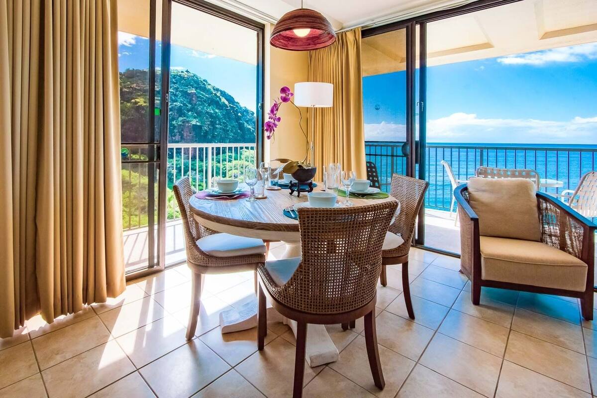 A balcony with a dining table set for four, wicker chairs, and a view of the ocean.