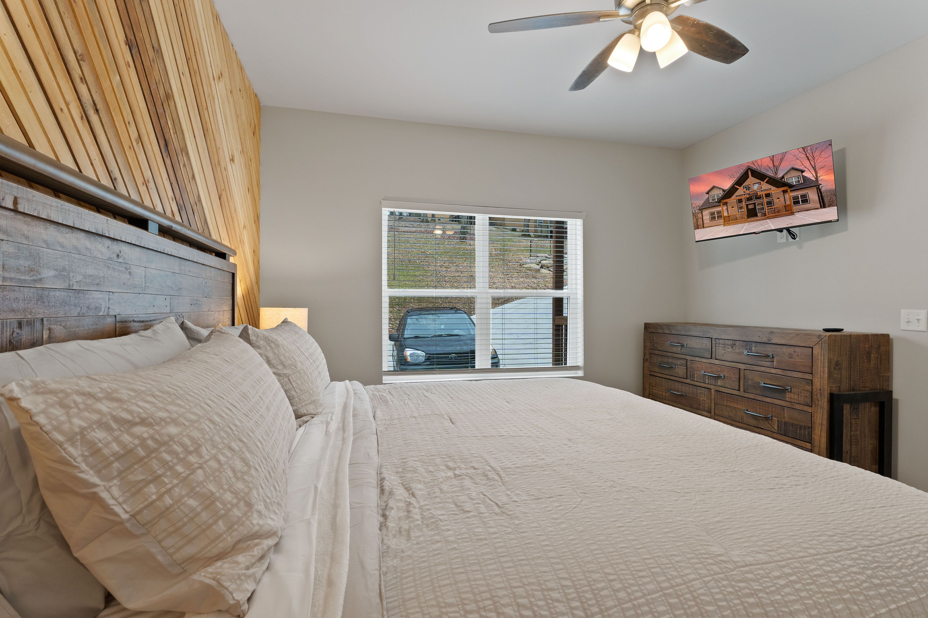 A bedroom with a bed, a dresser, a ceiling fan, a window with blinds, and a painting on the wall.