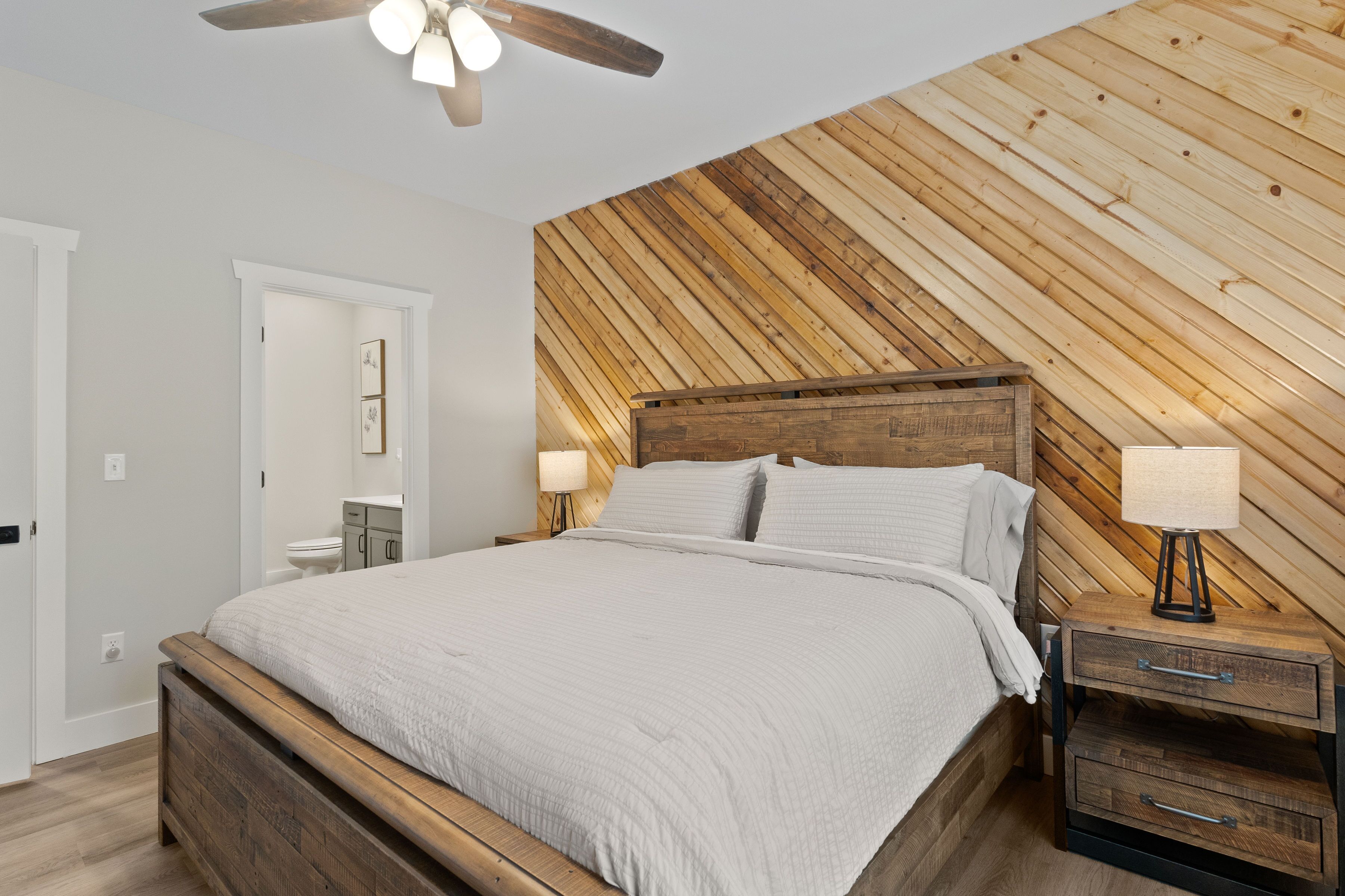 A bedroom with a wooden slat wall, a wooden bed frame, a nightstand with a lamp, and a ceiling fan.
