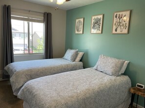 A room with two beds, a window with curtains, and three framed artworks on the wall.