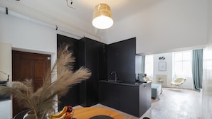 Cocina moderna con encimera negra, un lavamanos y una cafetera. Hay una zona de comedor con una mesa y sillas de madera, y un jarrón con plantas secas.