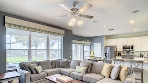 A spacious living room with a sectional sofa, a coffee table, and a dining area with chairs. There is a kitchen with white cabinets and a microwave.