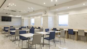 A modern conference room with white tables and chairs, a large flat-screen TV, and a city view through the windows.