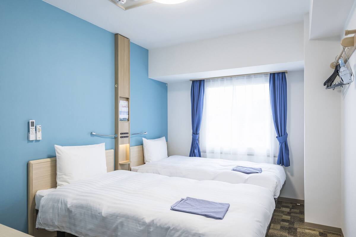 Een hotelkamer met twee bedden, een houten garderobe, een nachtkastje, een lamp en een raam met blauwe gordijnen.