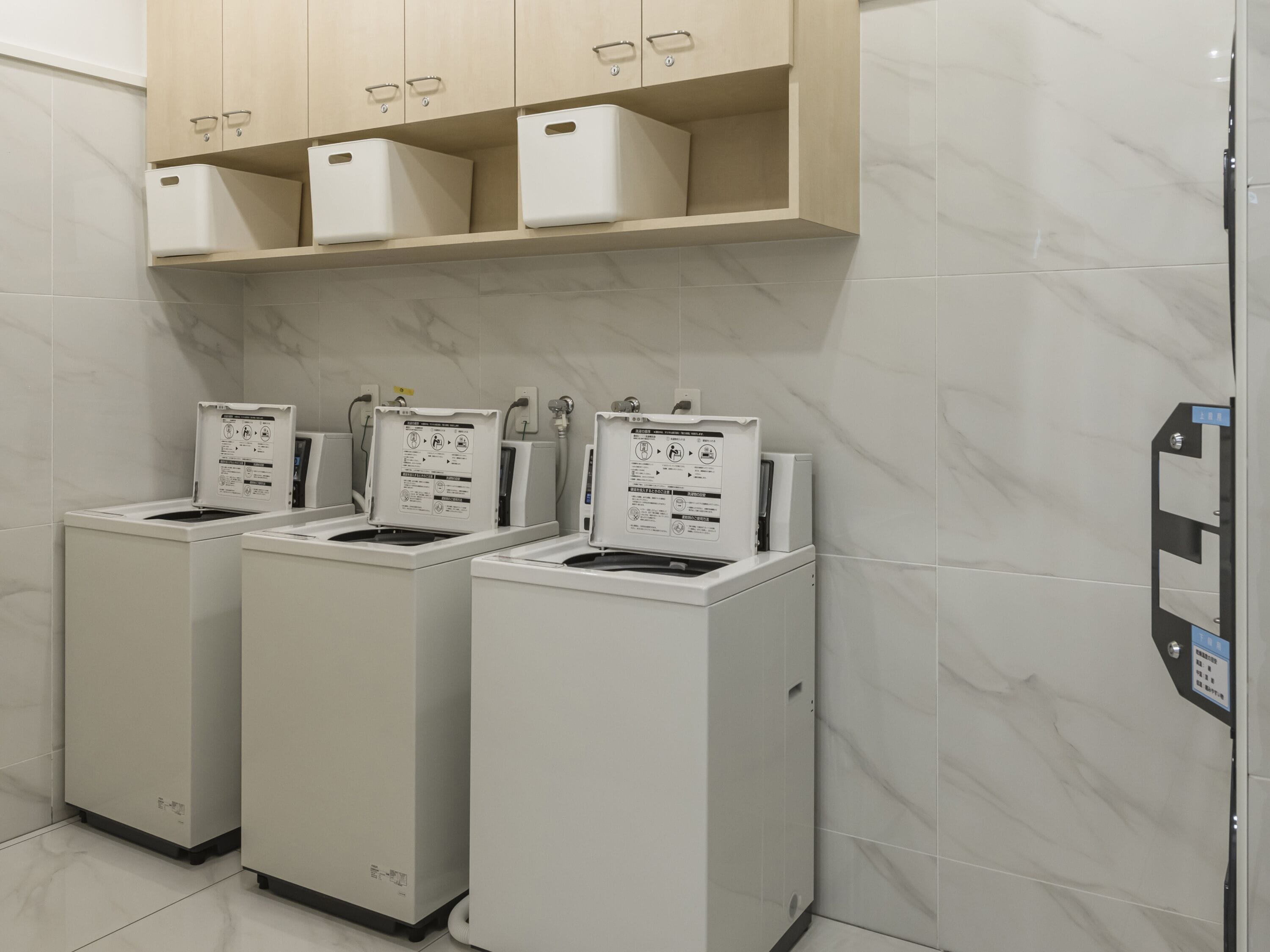 A laundry area with three stacked washing machines, a shelf above with storage boxes, and a wall-mounted control panel.