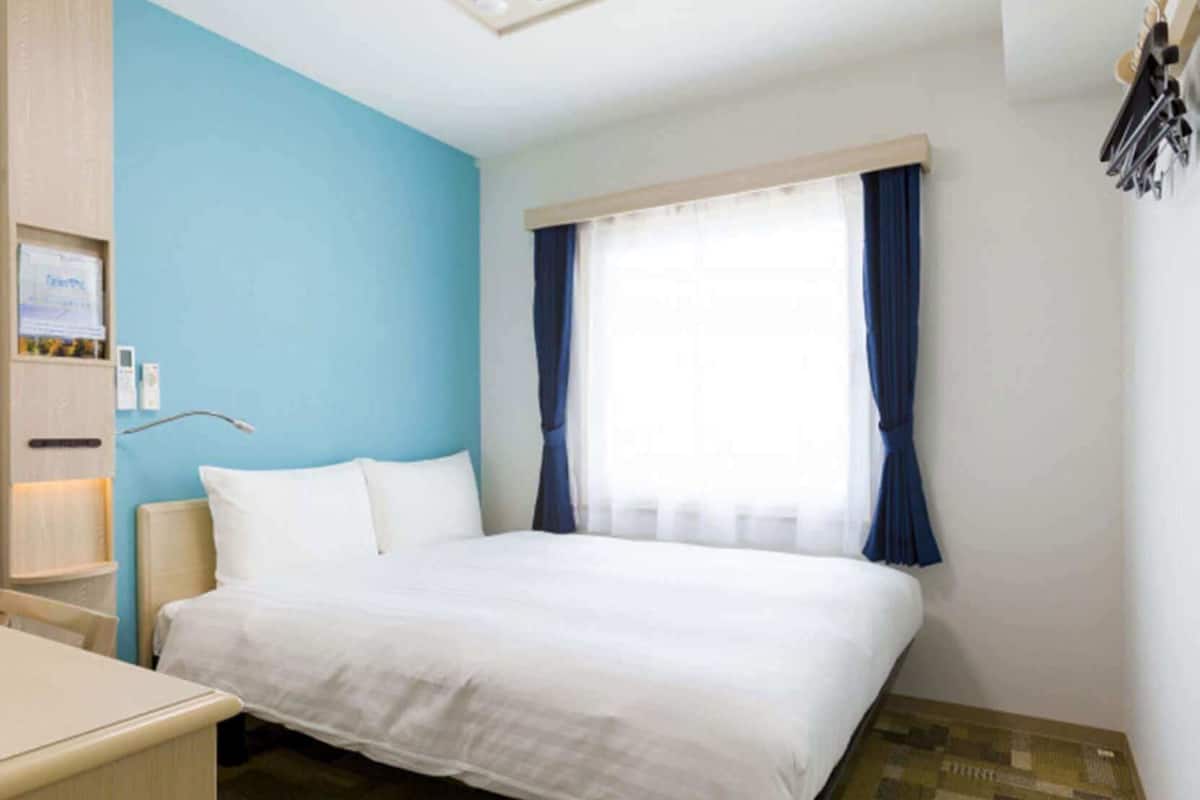 Hotelkamer met een bed, bureau, nachtkastje, raam met gordijnen en een wandplank met spullen.