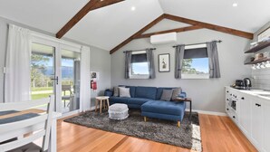 A cozy living room with a blue sofa, a small round ottoman, a wooden dining table with a chair, and a kitchen area with white cabinets and a stove. - Wakefields - an enchanting mountain view cottage (Mole Creek)