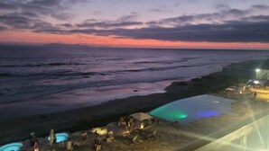 Un complexe hôtelier situé en bord de mer, doté d’une piscine, de chaises longues et offrant une vue sur l’océan au coucher du soleil.