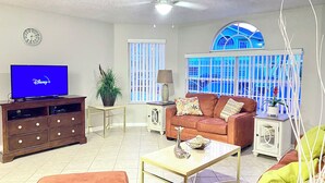 A living room with a television displaying the Disney+ logo, a brown entertainment unit, a red sofa with patterned cushions, a small table with a giraffe figurine, and a window with a view of a blue building. - Potter/Jedi vacation villa- Disney Parks 5 miles (Kissimmee)