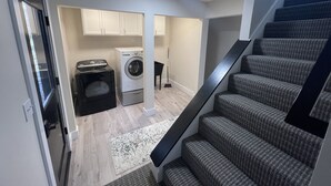Un espacio de lavandería pequeño con lavadora y secadora, una alfombra pequeña y una escalera con escalones cubiertos de moqueta gris.