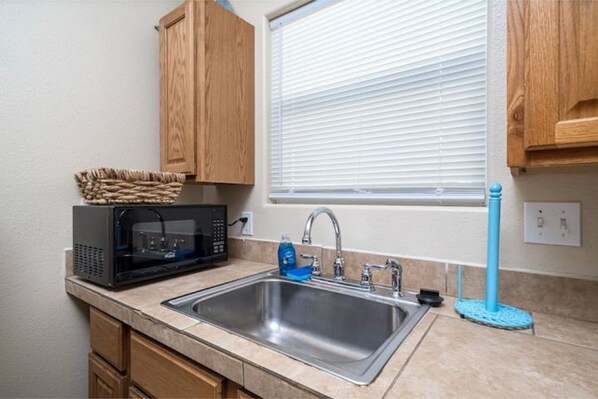 Una cocina compacta con un lavamanos de acero inoxidable, microondas, armarios de madera y una ventana con persianas.