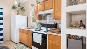 A compact kitchen with wooden cabinets, a white refrigerator, a stove, and a sink. There are decorative items on the shelves and a window above the sink.