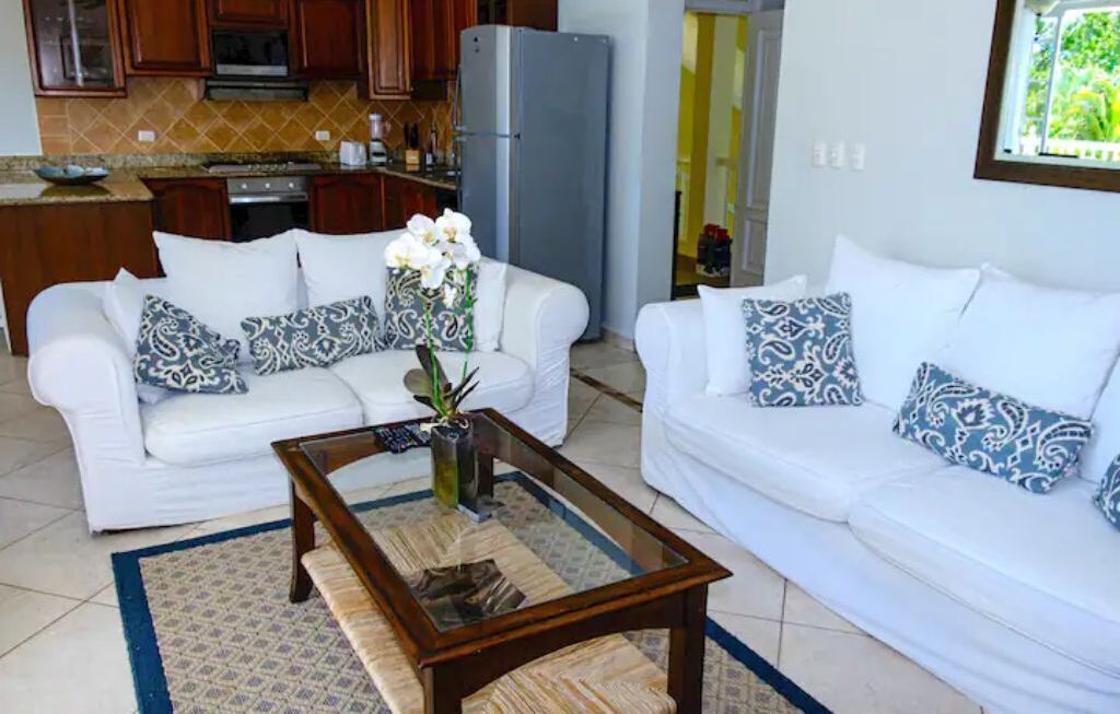 A living room with two white sofas, patterned cushions, a glass coffee table, and a kitchen in the background.
