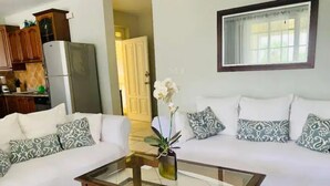 Ein Wohnzimmer mit Glastisch, weißen Sofas mit gemusterten Kissen, einer Küchenzeile mit Holzschränken und einem Kühlschrank sowie einem Fenster mit Vorhang.