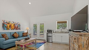 A spacious living room with a blue sofa, colorful rug, and a large painting. Adjacent is a kitchen with white cabinets, a microwave, and a large TV.