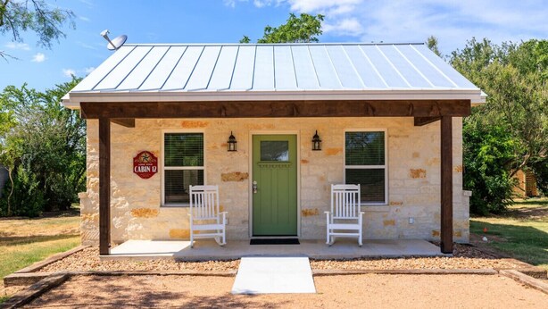 A small stone building with a green door, two white chairs outside, and a sign that reads "Cabin 12".