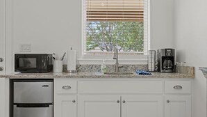A kitchen with white cabinets, a stainless steel refrigerator, a microwave, a coffee maker, and a window with blinds.