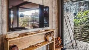 A television mounted on a wooden console with a rustic wall and a balcony with a view of greenery. - Sublime 1BR w Cenote | Big Gym | Pools | Jacuzzis (Tulum)