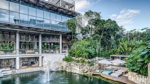 A modern building with large glass windows, a stone water feature, and outdoor seating areas surrounded by lush greenery. - Sublime 1BR w Cenote | Big Gym | Pools | Jacuzzis (Tulum)