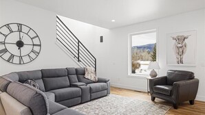 A modern living room with a grey sectional sofa, a black leather armchair, a large wall clock, a staircase, and a window with a mountain view. - Architectural Masterpiece Home with Mountain Views & Hot Tub (Red Lodge)
