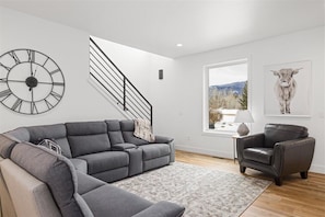 Una sala moderna con un sofá seccional gris, un sillón de cuero negro, un reloj de pared grande, una escalera y un ventanal con vista a la montaña.