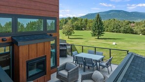 A modern outdoor patio with a fireplace, seating, and a view of a grassy field and mountains. - Architectural Masterpiece Home with Mountain Views & Hot Tub (Red Lodge)