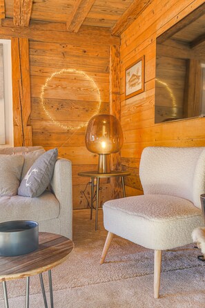A cozy room with wooden walls, a round table with a lamp, a sofa, and a chair. - Magnificent chalet 5 minutes from the center of Megève! (Megève)