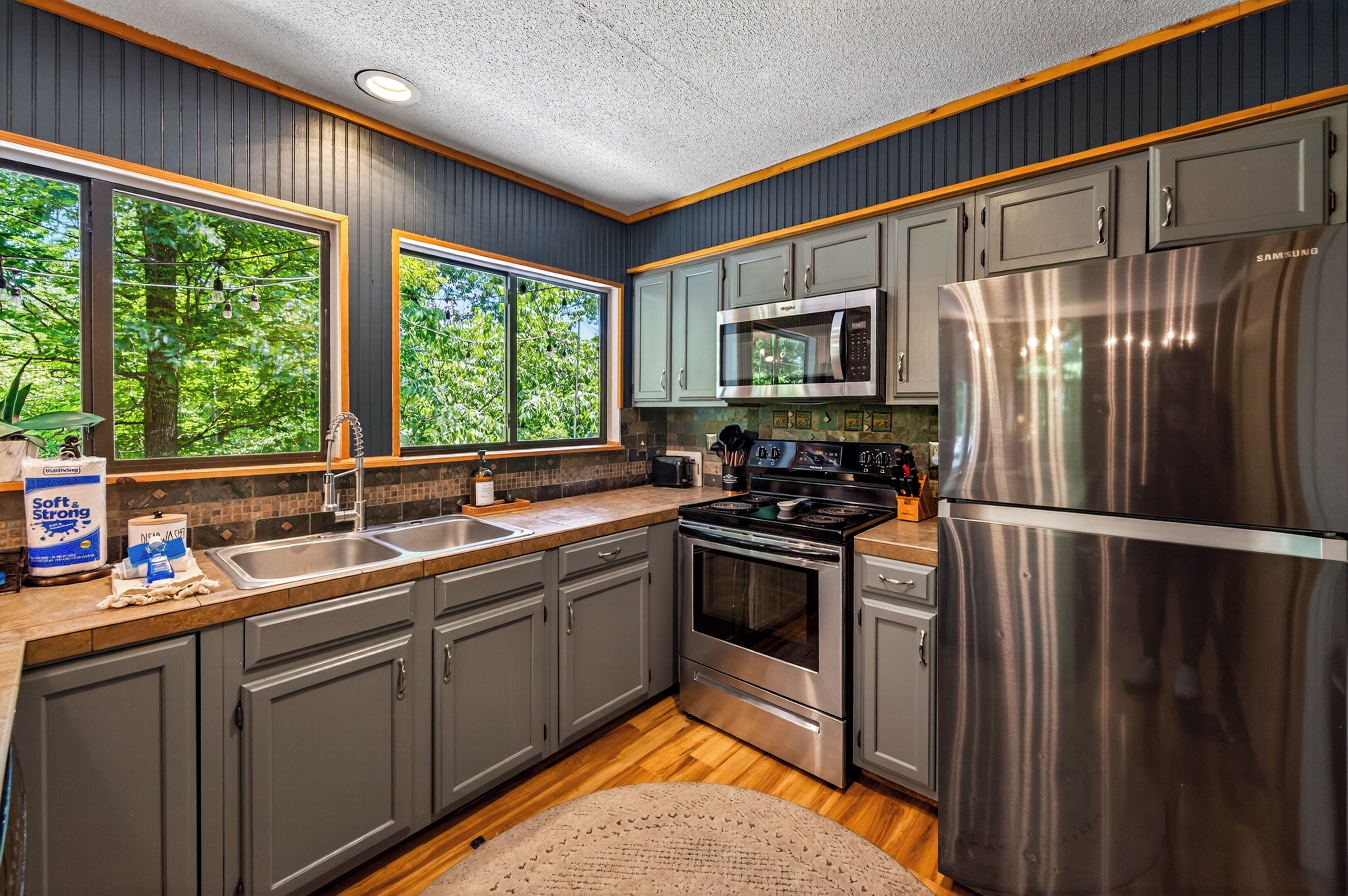 A modern kitchen with grey cabinets, stainless steel appliances, and large windows.