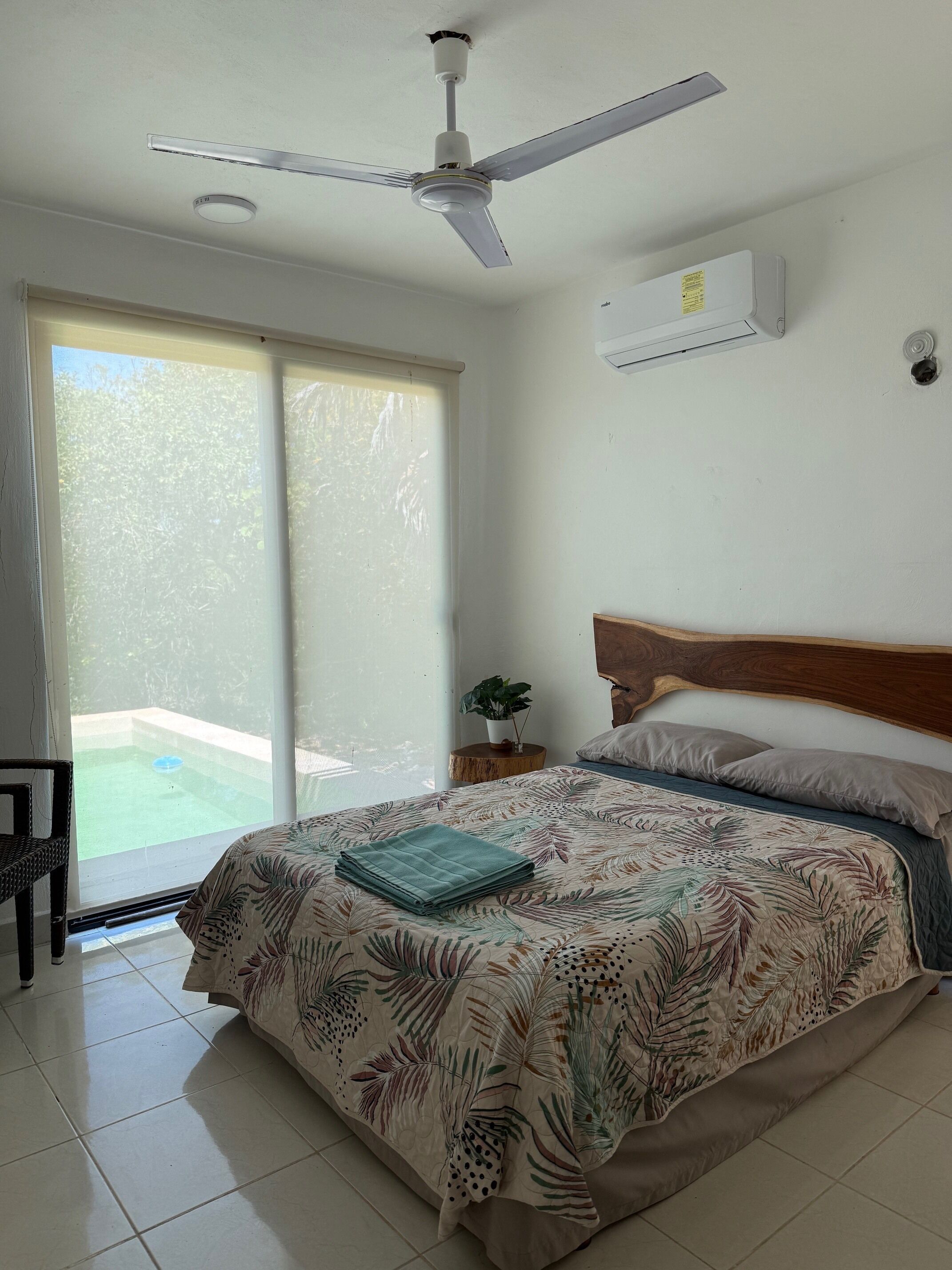 A bedroom with a bed, a ceiling fan, a window with a view of a pool, and a small bedside table with a plant.