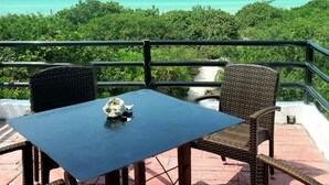 Un balcó amb cadires de vimet i una taula, amb vista al mar turquesa i vegetació densa.