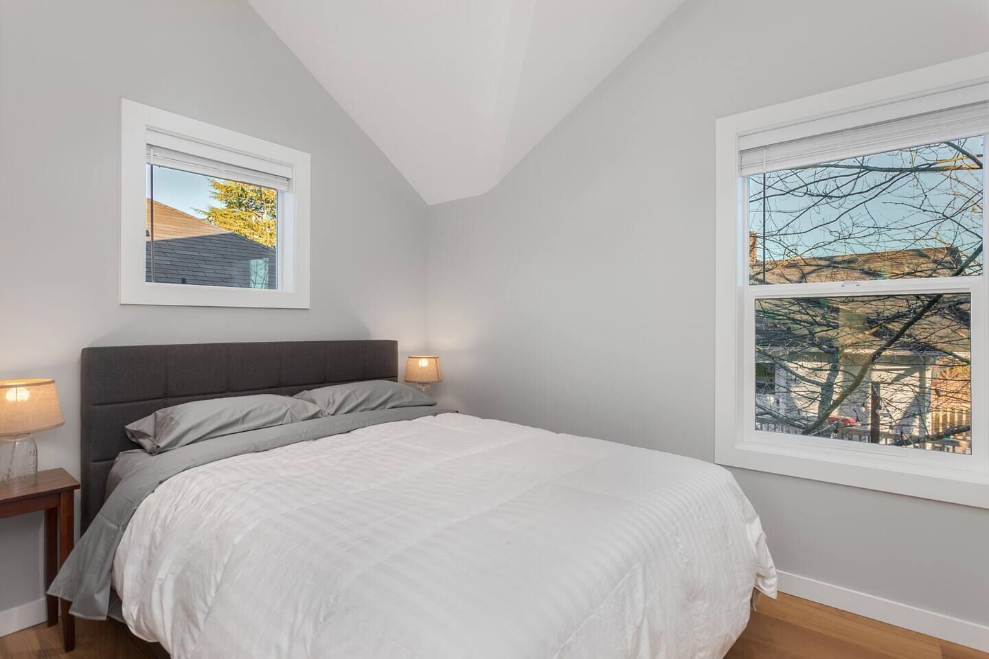 A bedroom with a bed, two bedside lamps, a framed picture on the wall, and a window with a view of trees and buildings.