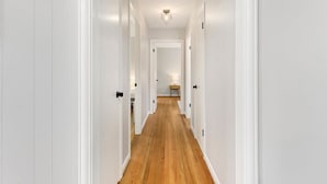 A narrow hallway with white walls and wooden flooring, leading to a room with a bed and a nightstand.