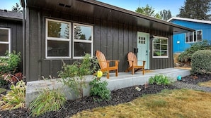 A modern house with a dark exterior, two windows, a door, and a porch with two wooden chairs.