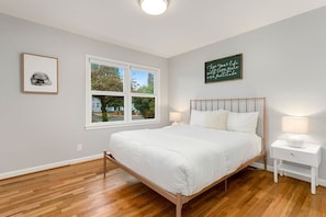 Un dormitorio con cama de madera, ropa de cama blanca, una ventana con vistas, un cuadro enmarcado y una frase motivacional en la pared.