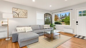 A modern living room with a grey sectional sofa, a glass coffee table, and a large window offering a view of trees and a house.